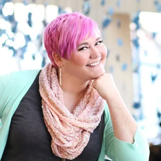 Dr. Debbie Burns with short pink hair, wearing a sparkly scarf and mint green cardigan, leaning on her hand in a bright, modern space. A happy client of Jackie Barker, reflecting the joyful and stress-free experience of working with Aligned Soul Design for website and marketing services.
