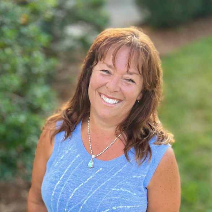 Terrie Hanscom wearing a light blue sleeveless top and a turquoise pendant necklace, standing outdoors with a blurred green background. A happy client of Jackie Barker, reflecting the ease and alignment found through Aligned Soul Design’s website and marketing services.