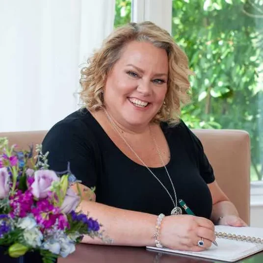 Kim Woods wearing a black top and silver jewelry, sitting at a table and writing in a notebook. A soulful design client of Jackie Barker, reflecting the ease and confidence gained from working with Aligned Soul Design for website design and digital marketing.