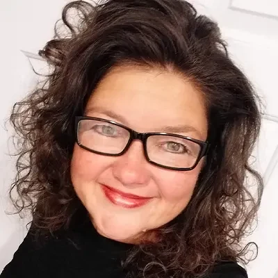 Michelle Cordy, wearing black-framed glasses and a black top, standing against a white background. A happy client of Jackie Barker, reflecting the confidence and clarity gained from working with Aligned Soul Design for website and marketing solutions.