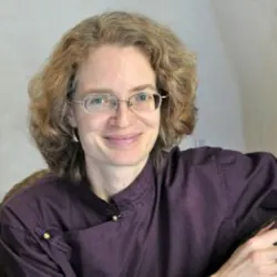 Andrea M Winn with curly light brown hair and glasses, wearing a deep purple blouse, sitting in a relaxed pose. A guest blogger contributing insights to Aligned Soul Design’s community of spiritual entrepreneurs.