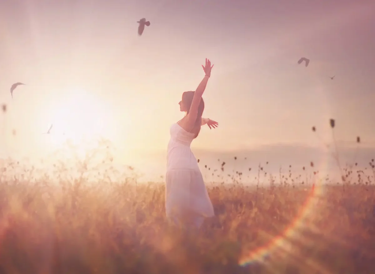 Girl standing in a sunlit field, surrounded by tall grass and wildflowers, looking peaceful.