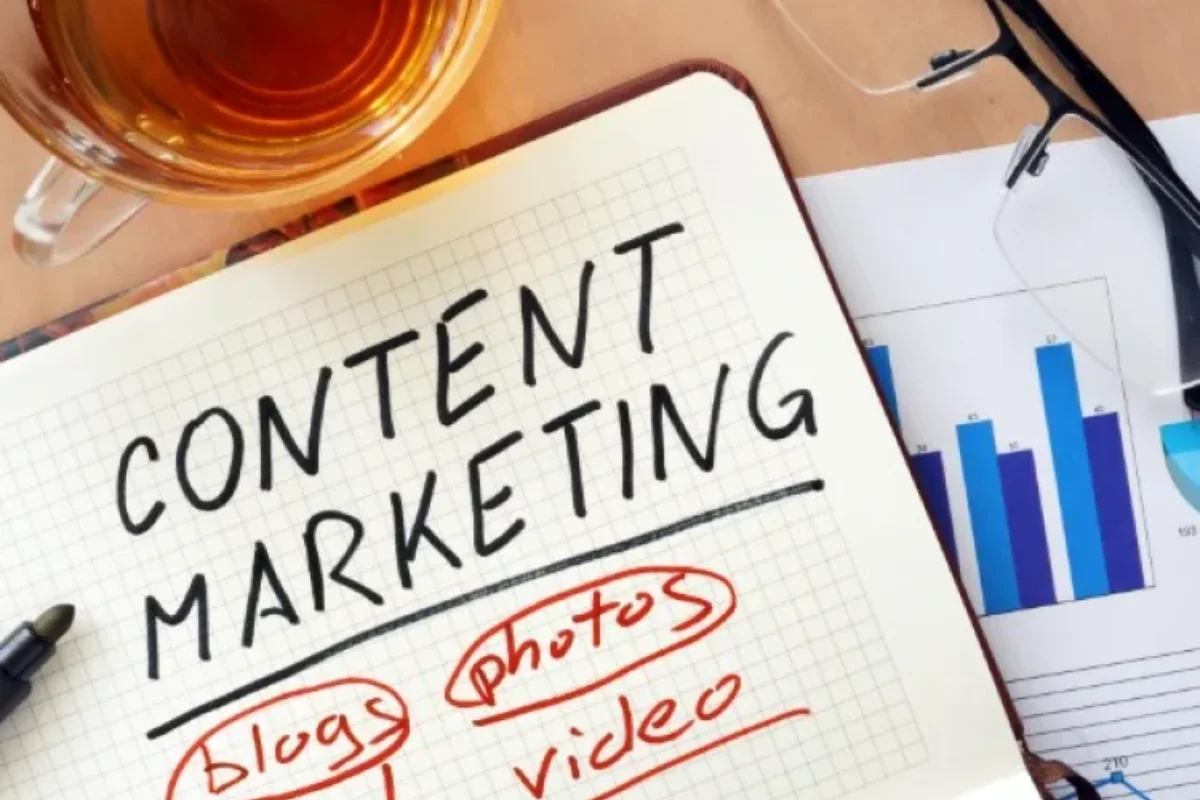 A notebook with "Content Marketing" written on it, surrounded by circles labeled "blogs," "photos," and "video." A cup of tea, glasses, and a graph are visible in the background.