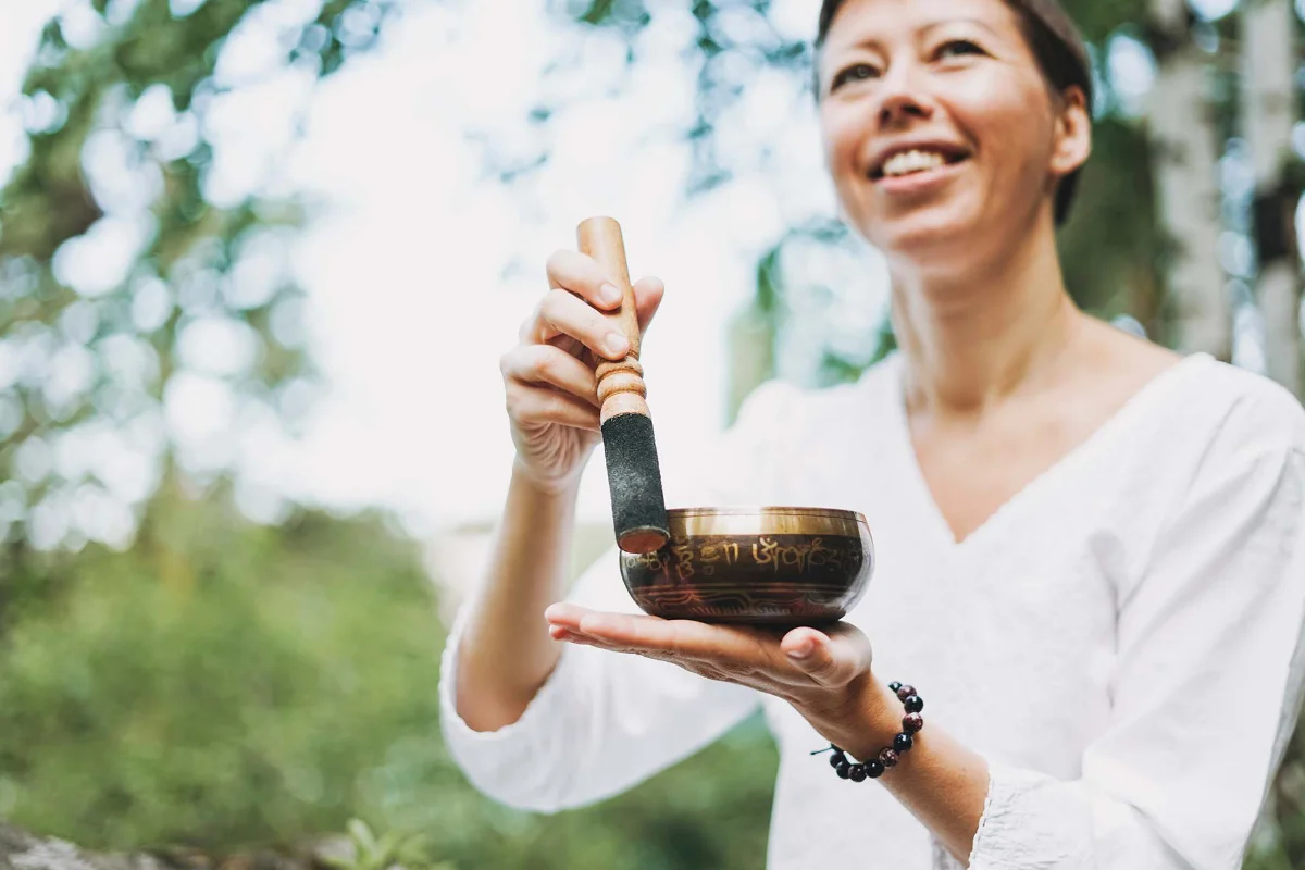 Person holding a Tibetan singing bowl and smiling outdoors, using a mallet to create sound vibrations.