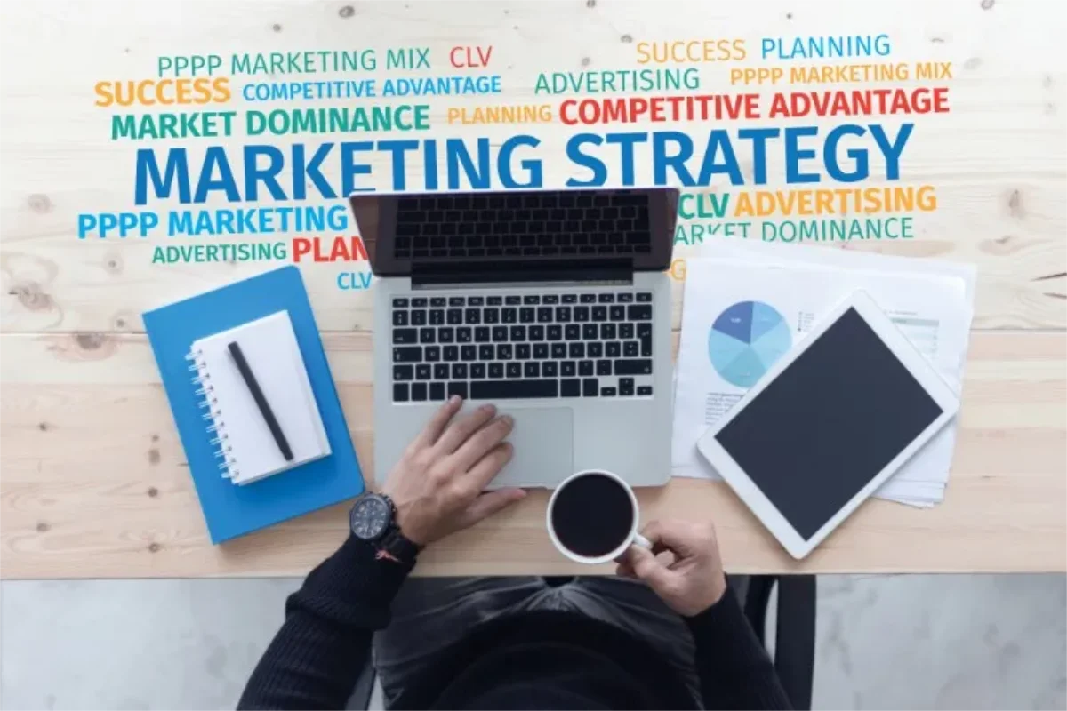Top-down view of a person working on a laptop with coffee, a notebook, and tablet, surrounded by marketing strategy keywords on the desk.