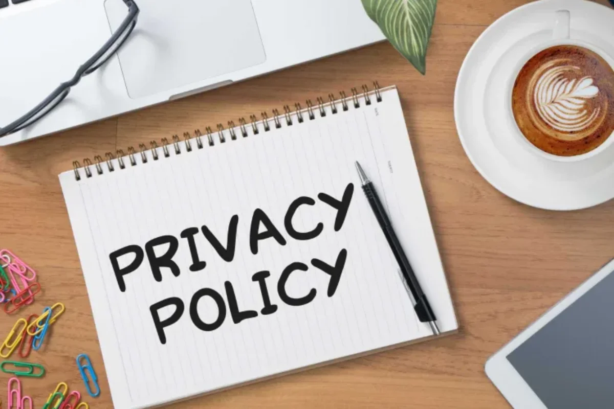 Notebook with the words 'Privacy Policy' written on it, placed on a wooden desk alongside a pen, a cup of coffee with latte art, colorful paper clips, and a tablet partially in view.
