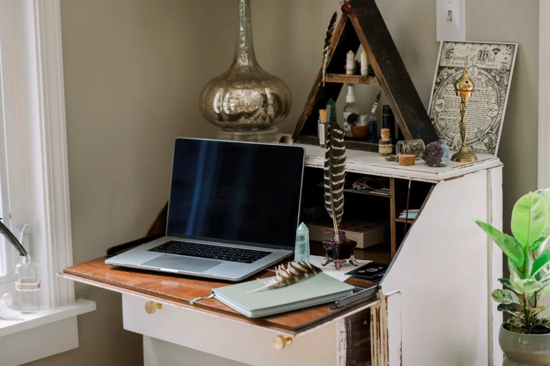 Rustic desk with an open laptop, notebook, feathers, crystal, and spiritual decor, including an altar and potted plant, bathed in natural light.