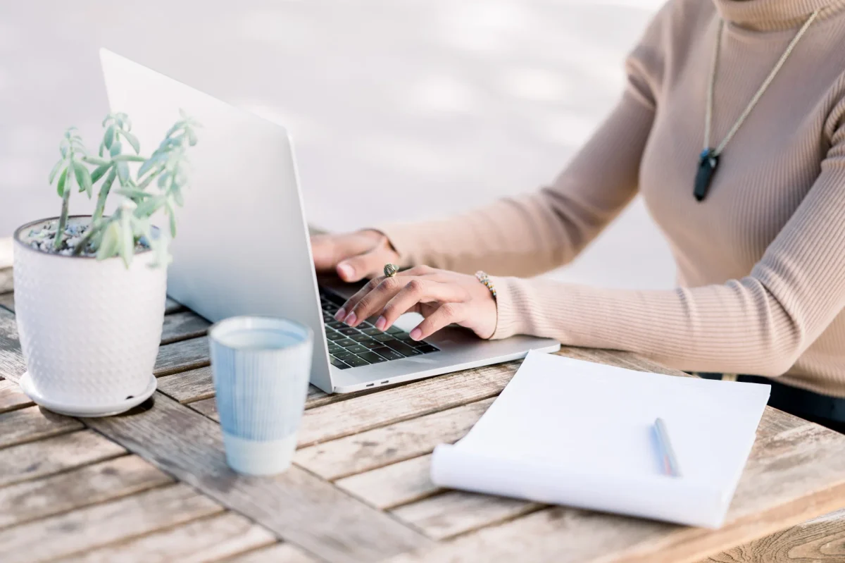 Close-up of a woman typing on a laptop at a wooden outdoor table, with a small potted plant, a ceramic cup, and a notepad with a pen nearby.