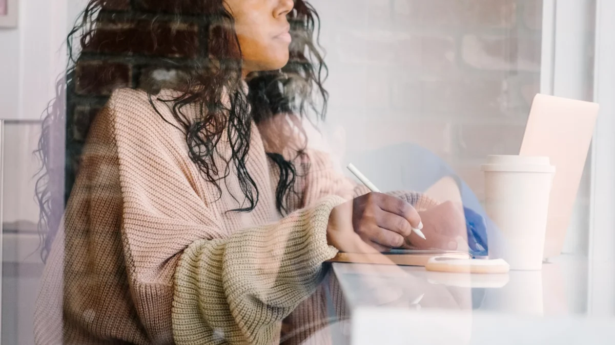 Woman writing in a notebook at a cafe with a laptop and coffee, representing planning with a website comparison checklist.
