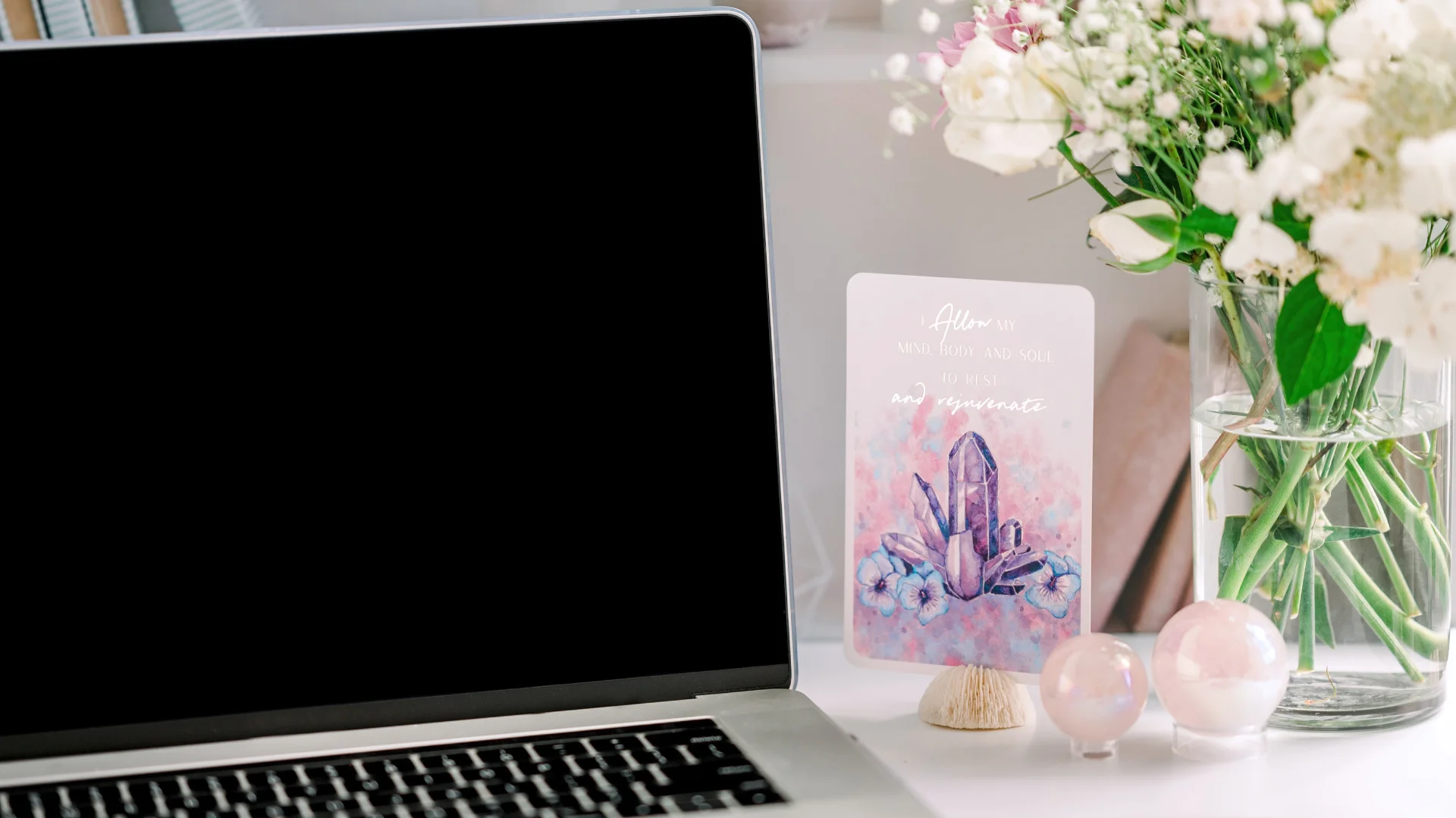 A laptop with a blank screen sits on a bright, modern desk next to a crystal-themed affirmation card that reads, “Allow my mind, body, and soul to rest and rejuvenate.” A vase of fresh flowers and two iridescent crystal spheres add a calming, spiritual touch.