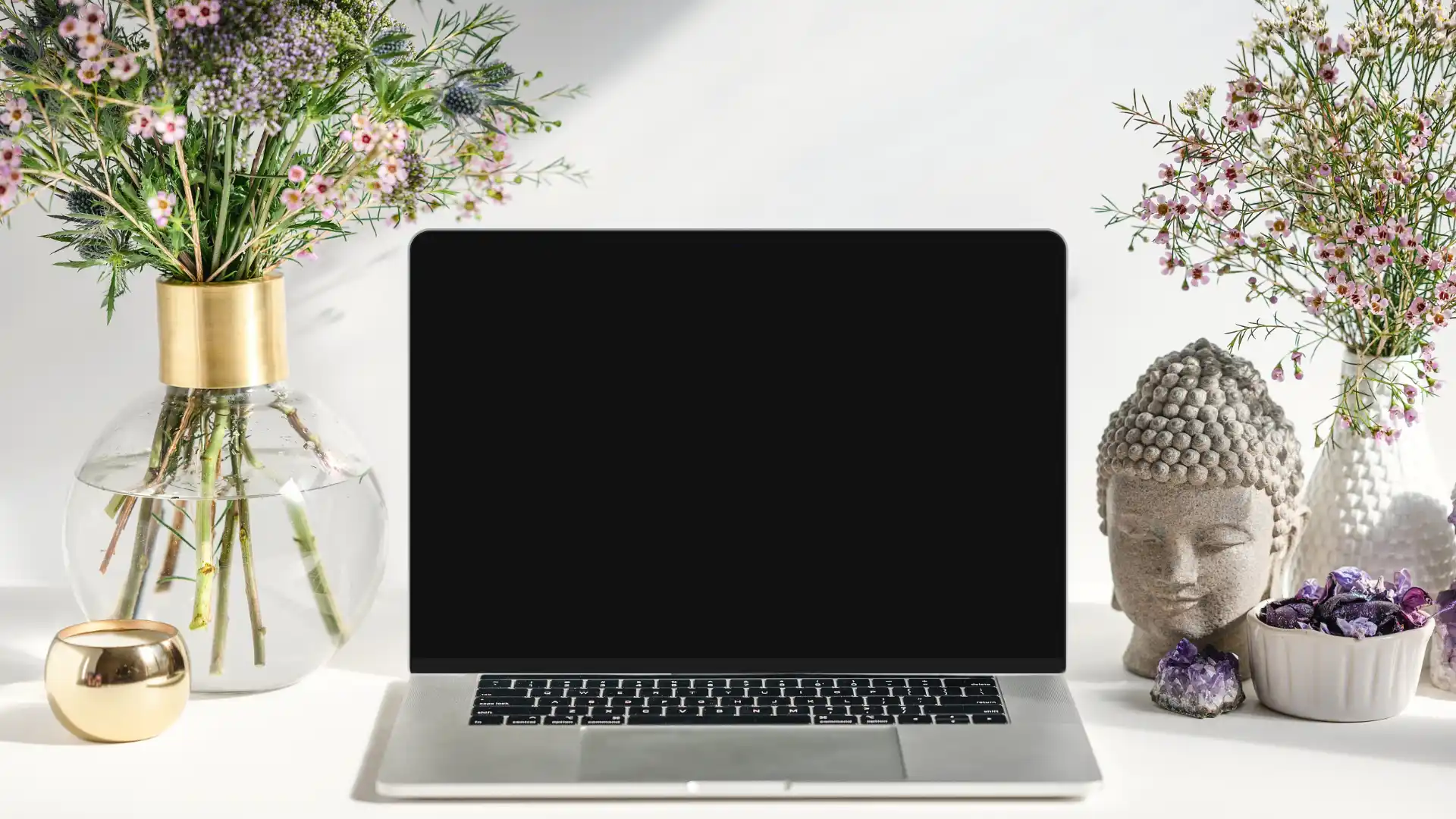 A laptop on a white desk surrounded by flowers, a Buddha statue, and crystals, representing balance and efficiency in website maintenance services.