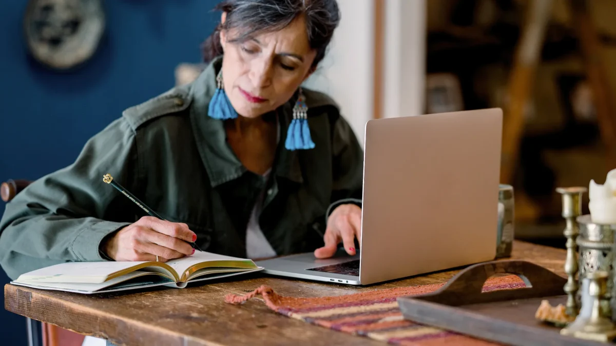 A woman sits at a wooden table, writing in a notebook while working on a laptop. She appears focused, suggesting she is brainstorming or writing content. The setting is warm and creative, with decorative elements in the background.