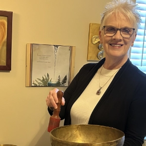 Dr. Betsy Goggan, a smiling older woman with short white hair and glasses, stands in a peaceful room holding a Tibetan singing bowl. She wears a black cardigan over a white top, with framed artwork and soft lighting in the background—grateful for the ease of her website pay monthly plan from Aligned Soul Design.