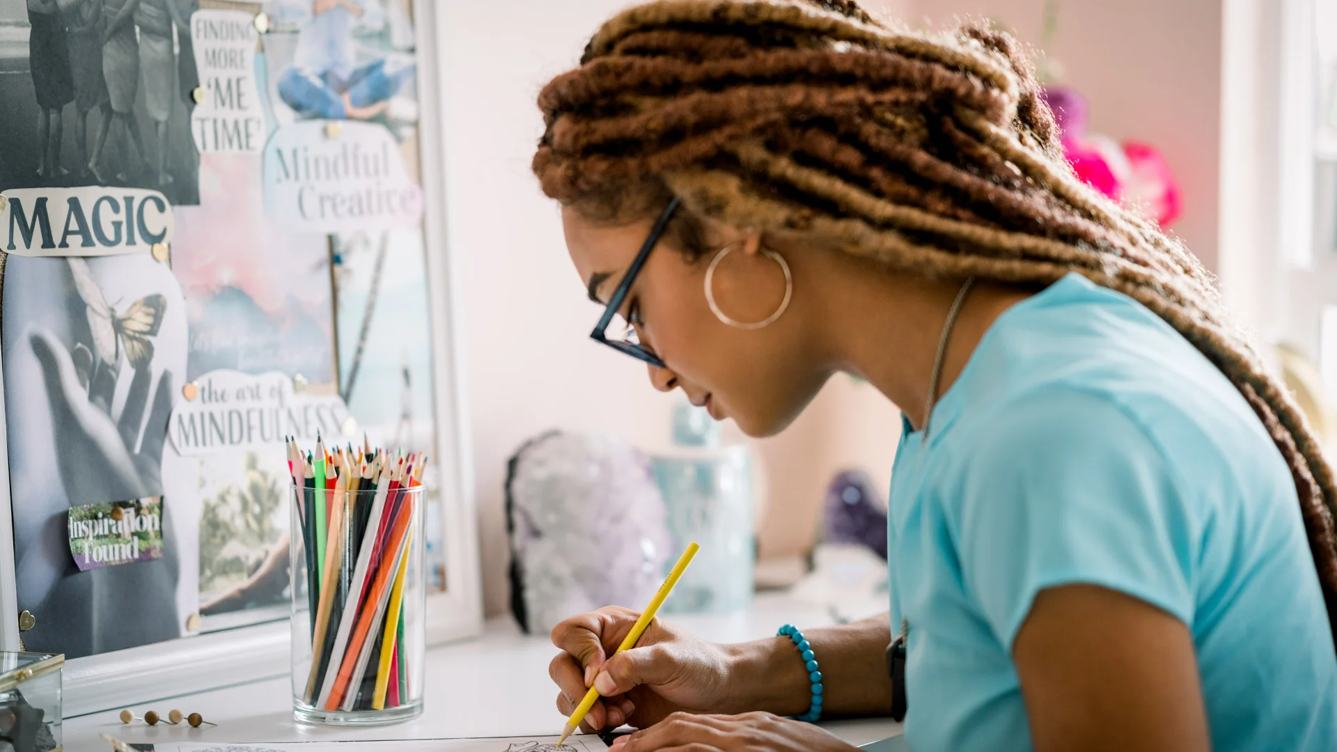 Woman sketching at a desk with colored pencils, crystals, and a vision board that says “Magic,” representing spiritual graphic design and creativity.