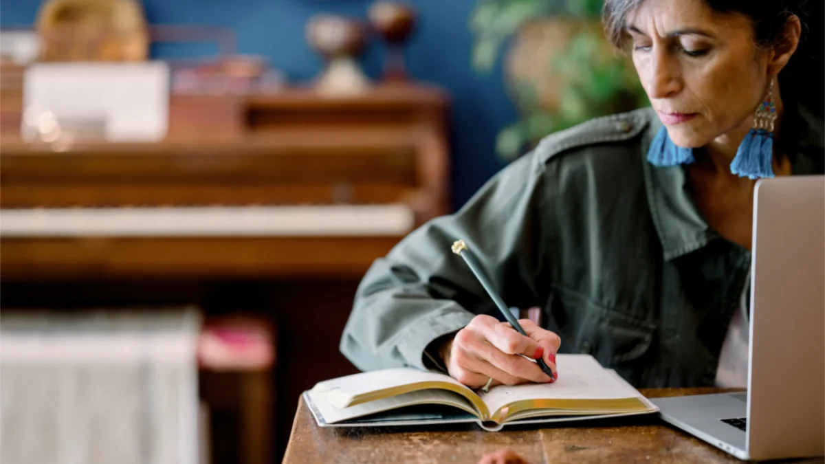 Woman writing in a notebook while working on a laptop at a wooden table, with a piano blurred in the background.