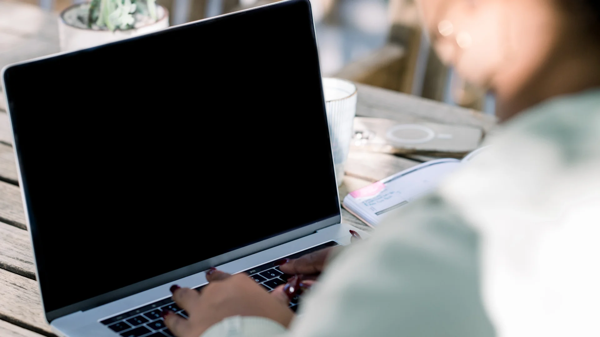 Woman working on a laptop outdoors at a wooden table, with a notebook and coffee nearby, representing focused, intentional online work.